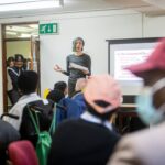 A diverse group attending a community meeting with a presenter speaking in front of a screen.