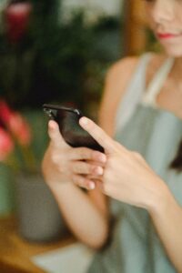 Unrecognizable female wearing apron text messaging on cellphone while standing near blurred flowers in vase in floristry store during work
